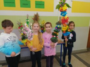 Od lewej strony stoją Szymon, Maja, Marietta i Basia. Dzieci prezentują swoje prace. Maja dumnie pokazuje swoją palmę ozdobioną różami i piórkami. Basia mocno trzyma swoją ogromną palmę wykonaną wraz z rodzicami. Palma ozdobiona bibułą oraz kolorową krepiną zdobyła I miejsce.